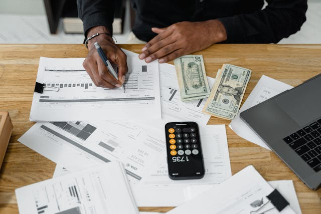 A person analyzes financial papers at a desk, surrounded by graphs, banknotes, a calculator, and a laptop.