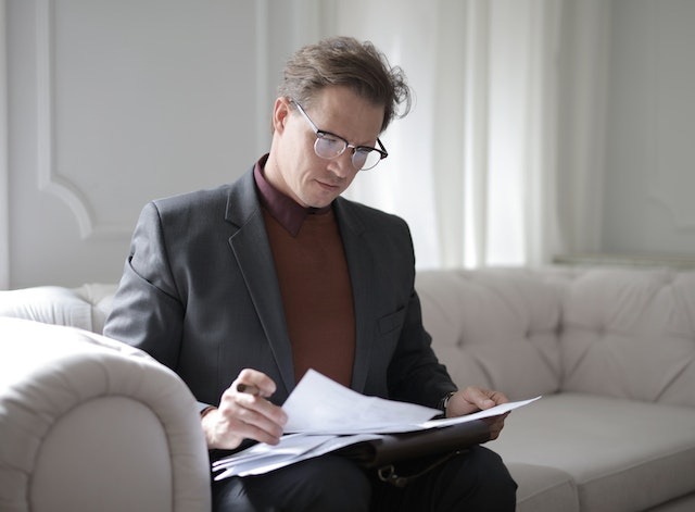 A man in glasses and a suit sits on a beige couch, focused on reading papers.